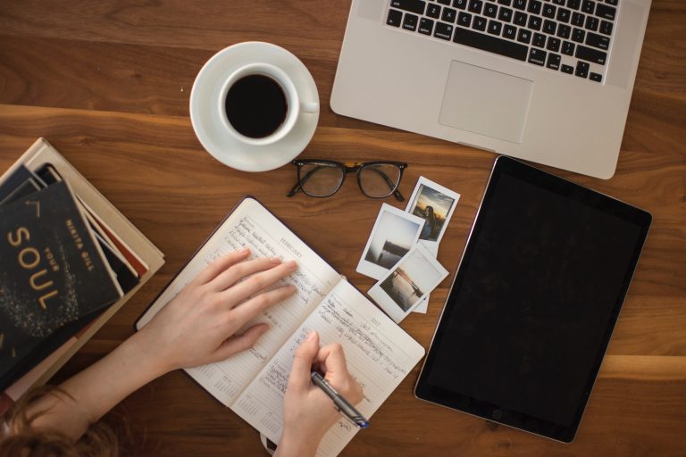 Person writes text in a notebook on a wooden table with her laptop in front
