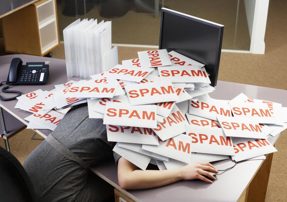 A woman in a grey dress sitting at the office desk, holding a computer mouse. There is a telephone, file cases, and a computer screen on the desk. The woman laid her upper body on the desk and covered herself with a huge number of white sheets of paper that have red letters on them saying "spam."