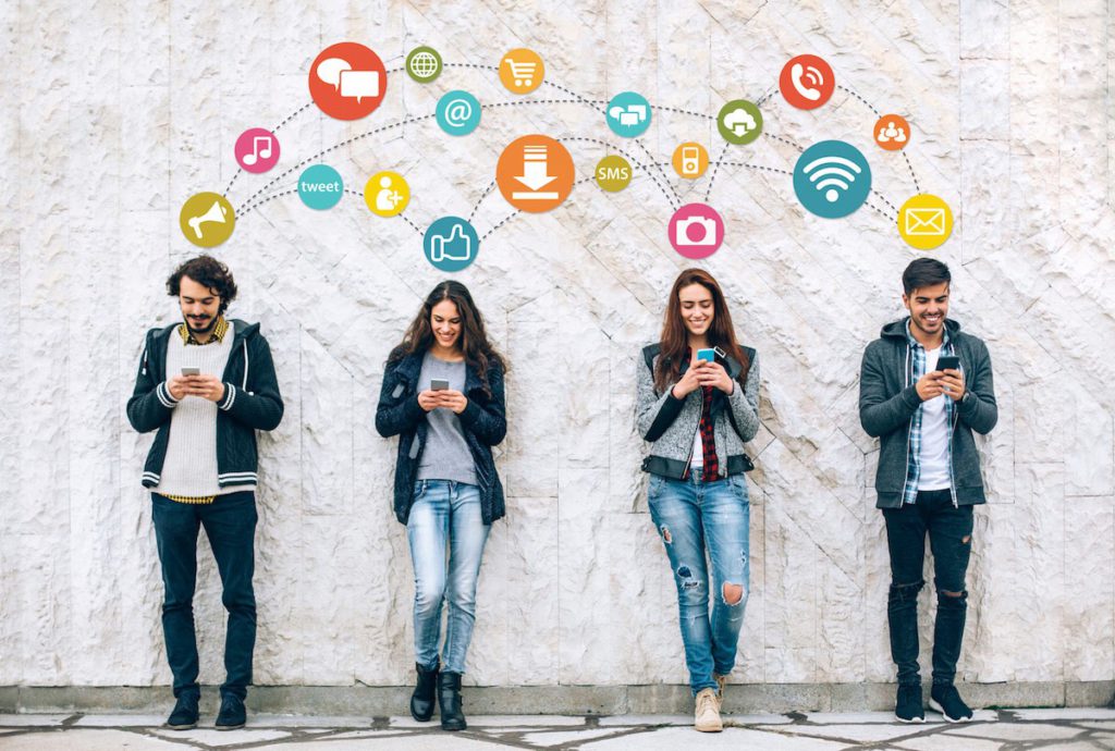 Four people standing against the wall and looking at their phones. The icons representing different activities on phone and social media are standing above them.